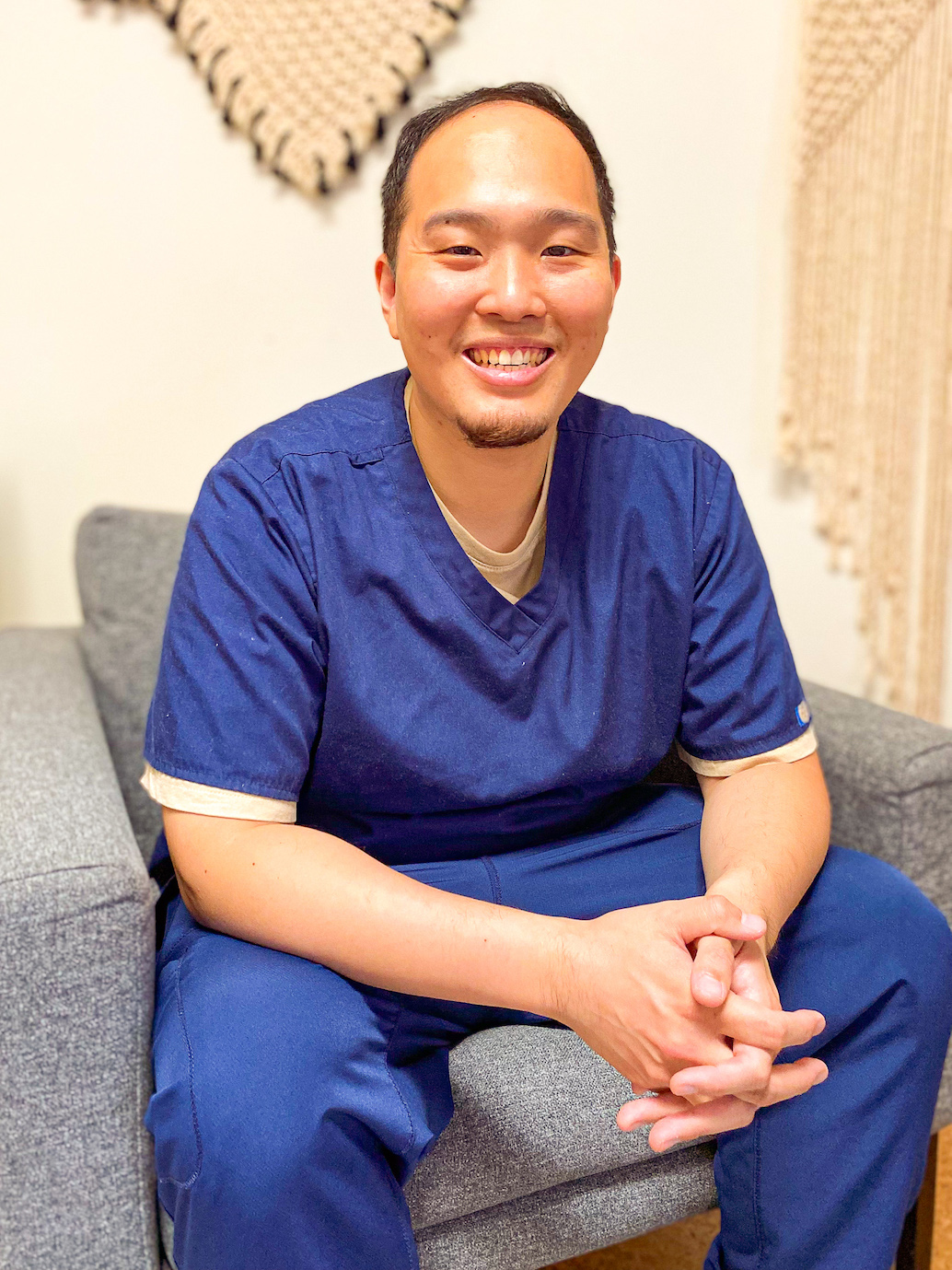 Doctor of acupuncture and traditional Chinese medicine looking at the camera sitting in grey chair and wearing blue scrubs.