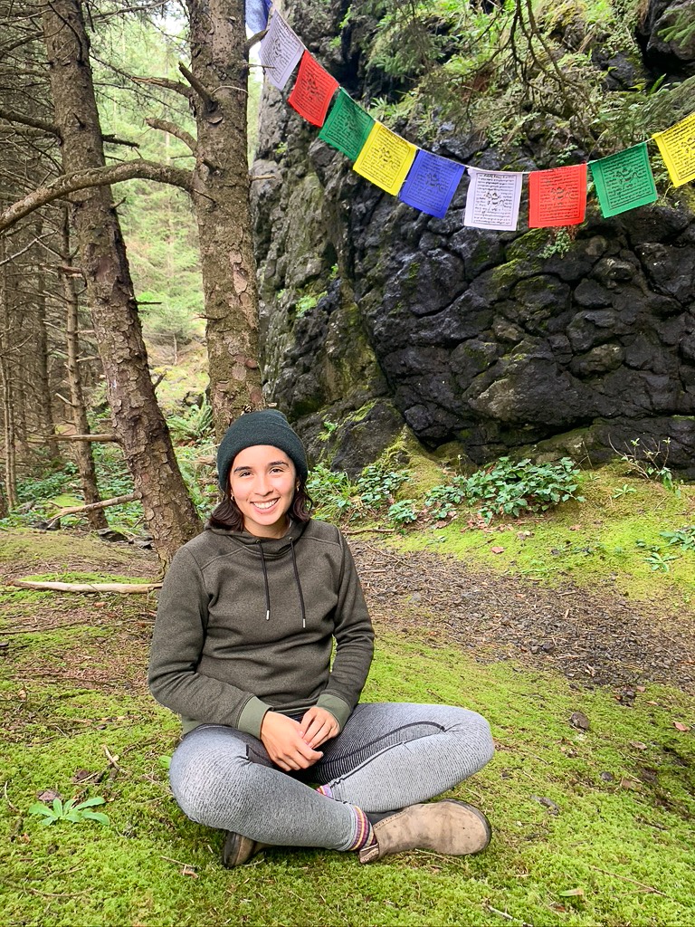 Woman sitting in on a plot of moss in a forest with Tibetan prayer flags in the background.